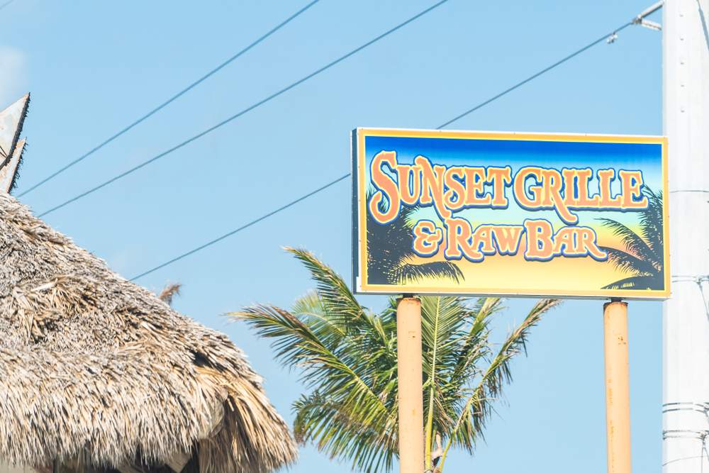 The outdoor sign for Sunset Grille stands outside the straw tiki hut roof of one of the best restaurants in Marathon, with a palm tree in the background.
