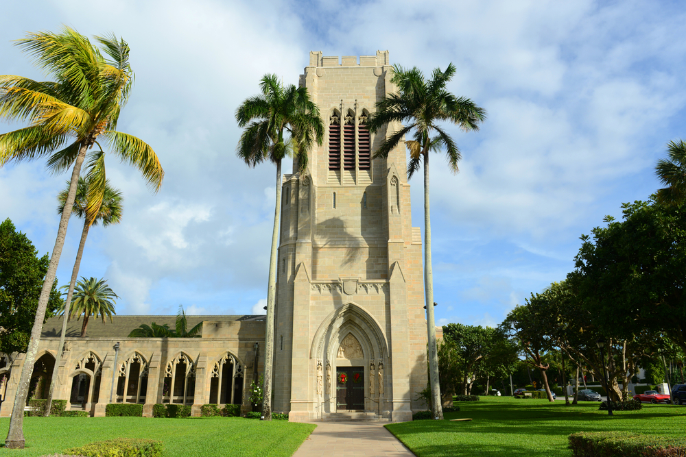 A large stone Gothic Revival church surrounded by grass and palm trees on a sunny day