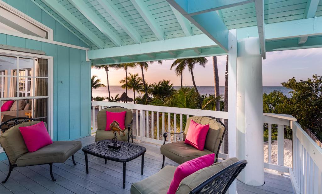 The private balcony of a room at a hotel right on the beach. It has chairs, a blue ceiling, white  posts, and views of the ocean and palm trees