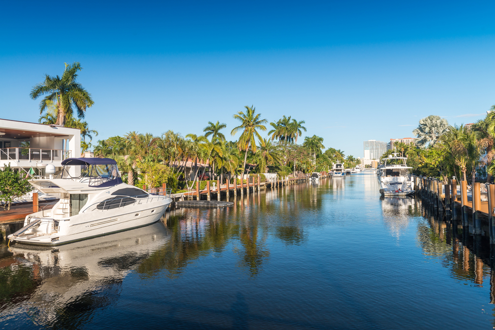 Boats sit docked outside homes along the palm-lined canals in Fort Lauderdale, FL on a sunny afternoon.