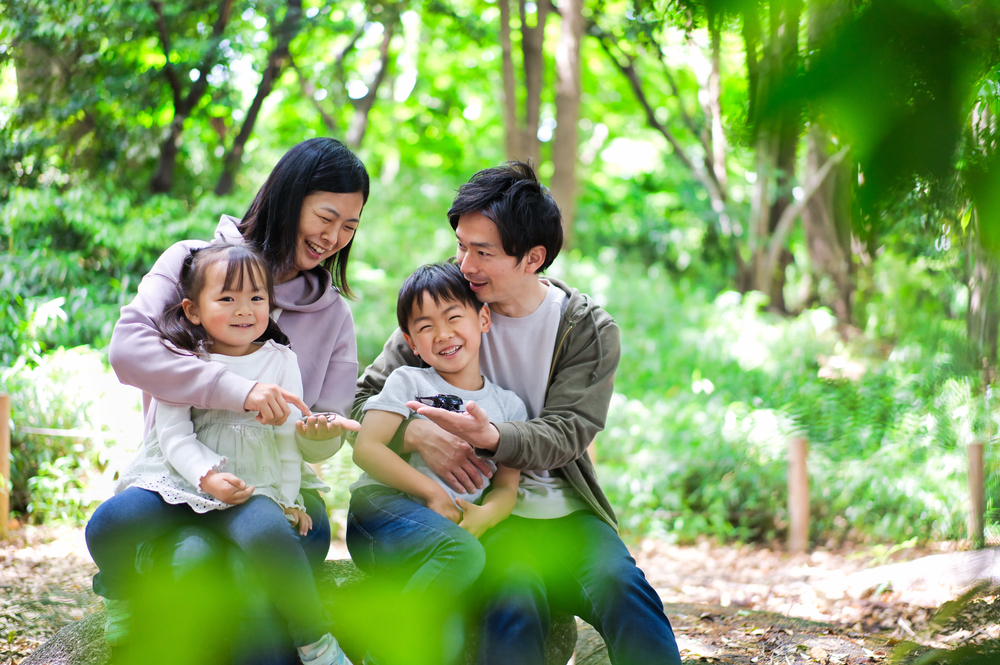An Asian family with a mom, dad, a young boy, and a young girl. They are in a wooded aviary and they have butterflies that have landed on their hands in Miami with kids
