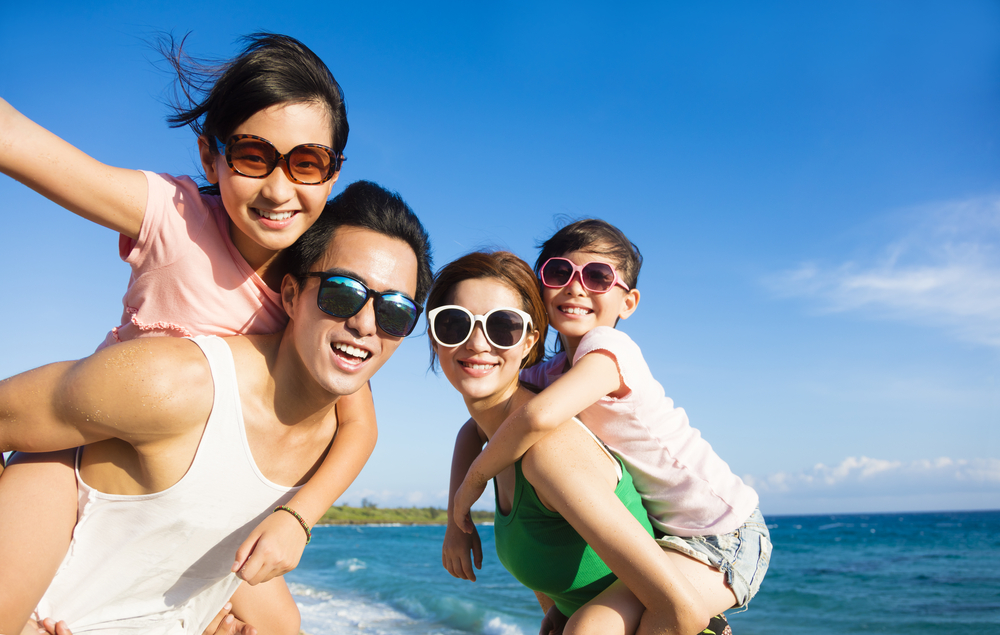 An Asian family with a mom, dad, and two young girls riding piggyback on their parents backs at the beach. They are all wearing large sunglasses in Miami with kids