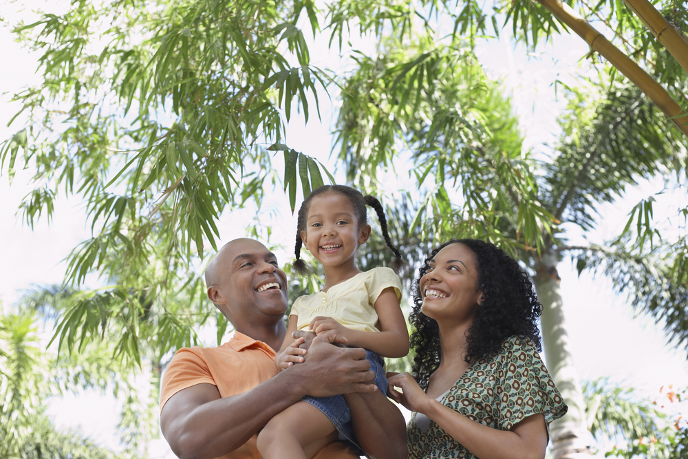 A Black family with a mom, dad, and young girl, smiling underneath palm trees in a park