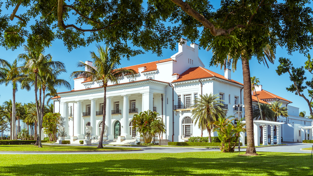 A white historic mansion surrounded by a lawn with palm trees that is now a museum