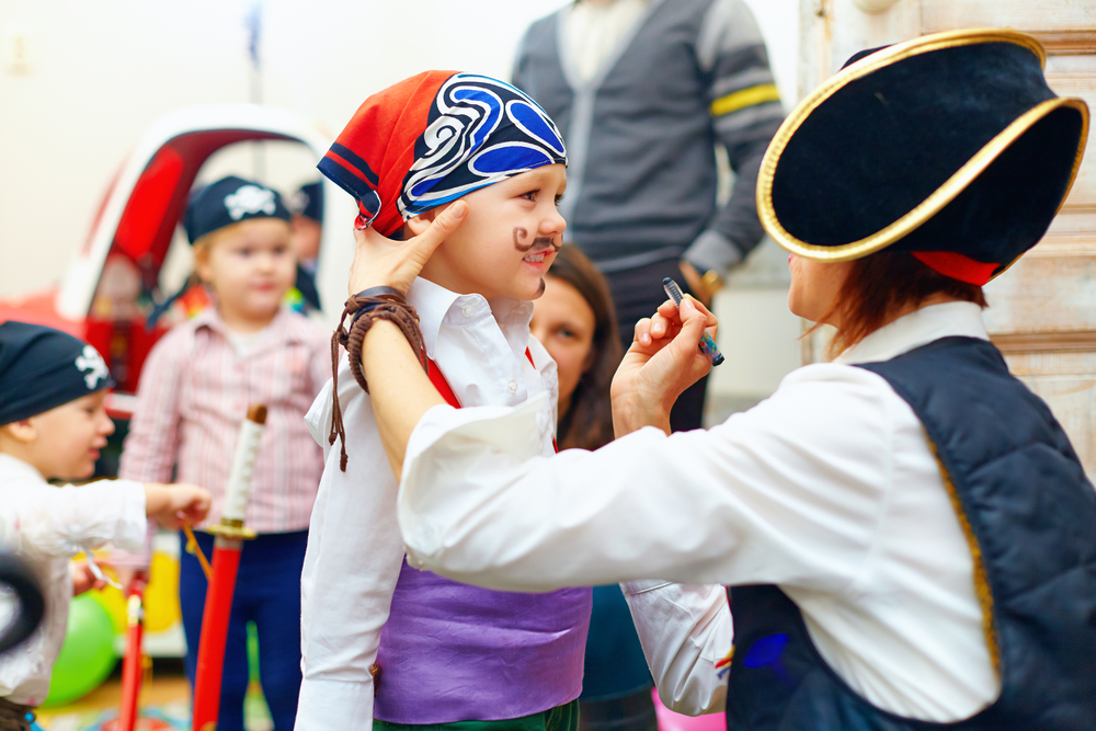 A young child wearing a pirate scarf and getting a mustache painted on their face by someone dressed as a pirate