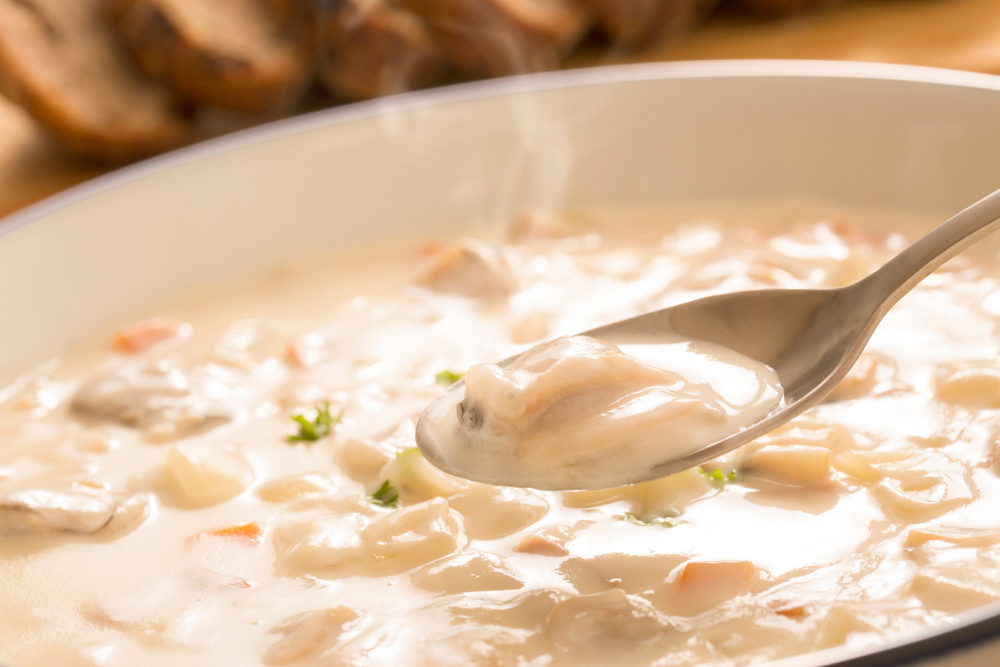 A spoon with pieces of clam meat lifts out of a bowl of warm, creamy clam chowder.