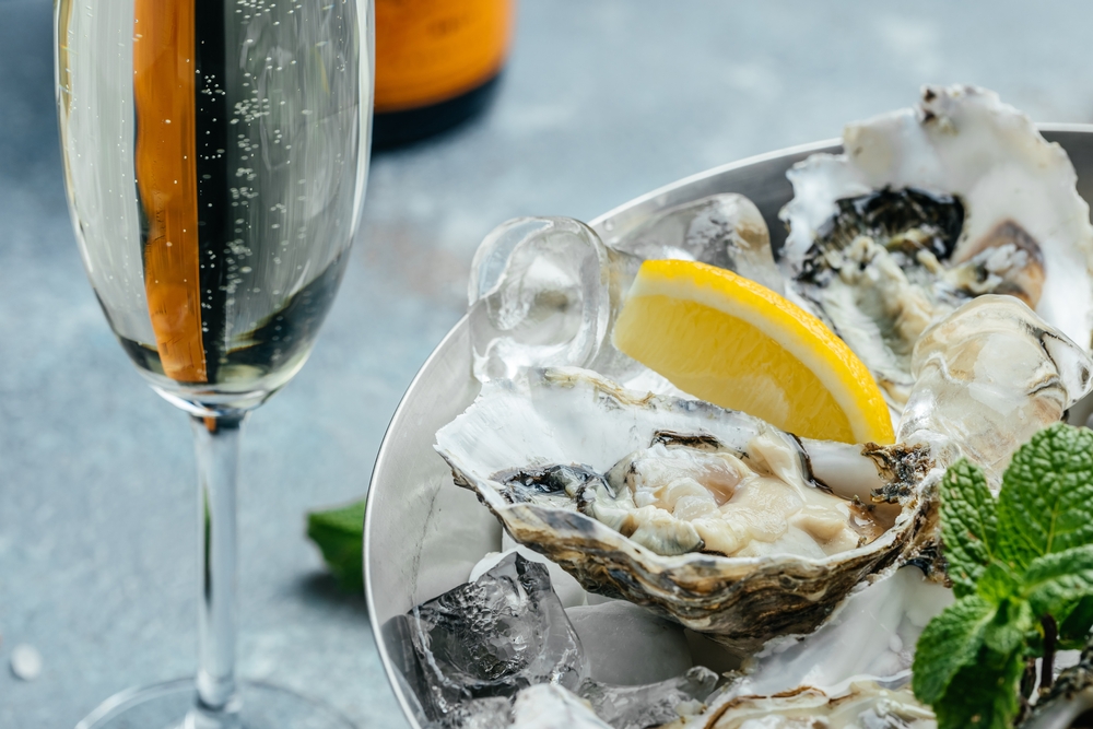 A glass of champagne stands next to an oyster and lemon wedge sitting on ice.
