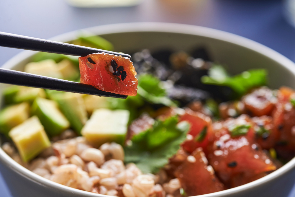 Two chop sticks hold a piece of raw tuna, pulled from a poke bowl with rice and avocado, much like the one served at USS Nemo, one of the best seafood restaurants in Naples.