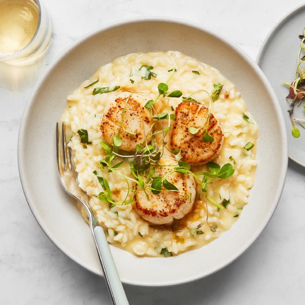 Three scallops sit on top of a creamy risotto, topped with green garnish, on a white plate, like the one served at Real Seafood Company, one of the best seafood restaurants in Naples.