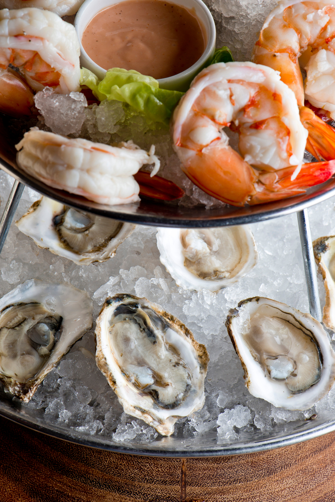 A two-tiered metal vessel holds shrimp and oysters on beds of ice, with a dipping sauce in the middle.