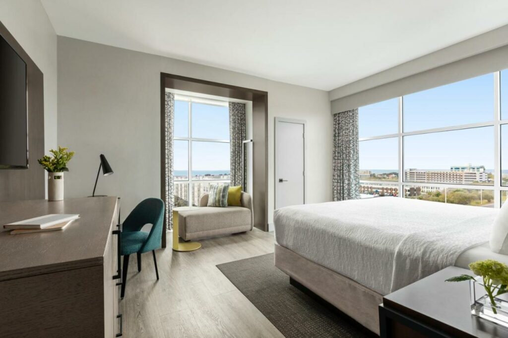 A room in a hotel that overlooks a cityscape and the ocean with large windows and neutral décor