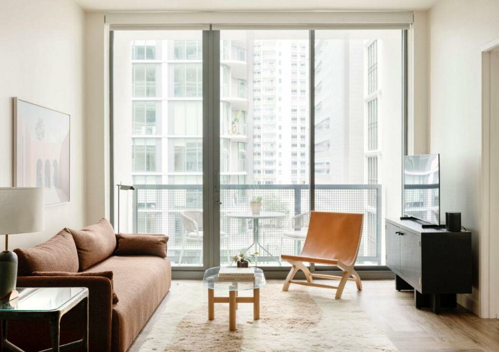 A neutral toned sitting area in a hotel room that has a balcony and looks out at the skyline of a city. 