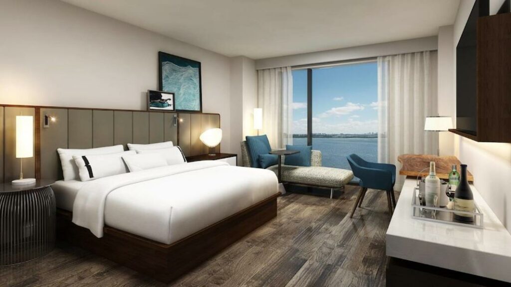 A modern hotel room with a view of the  ocean out of a large window that takes up the back wall of a hotel room