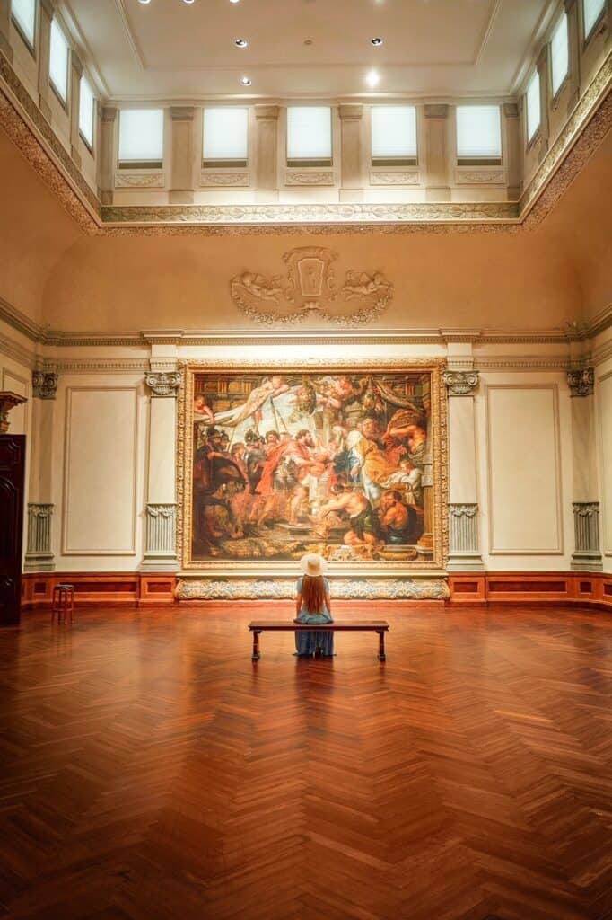 A woman with long hair sits on a bench, surrounded by hardwood flooring, looking at a large painting inside the Ringling Museum in Florida.