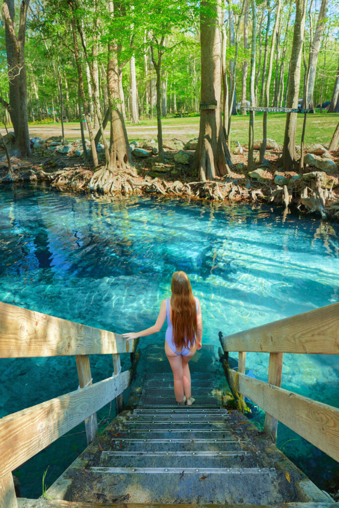 Woman with long hair walks down wooden stairs into bright blue spring water.