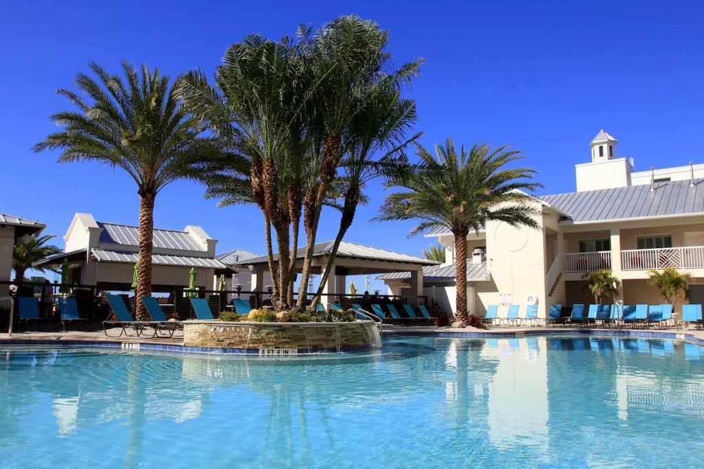 Hotel pool with palm trees and building in the background. The article is about the best beach resorts In Clearwater. 