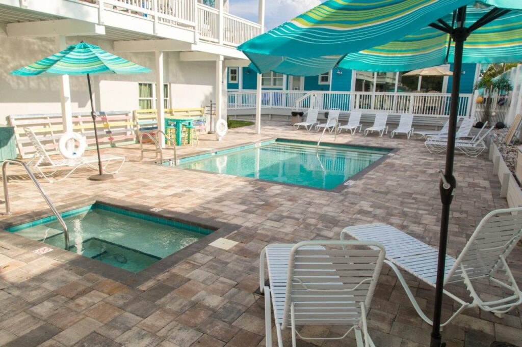 The pool area of a retro bed and breakfast that has a pool, a hot tub, blue and green stripped umbrellas, and white sun loungers. 