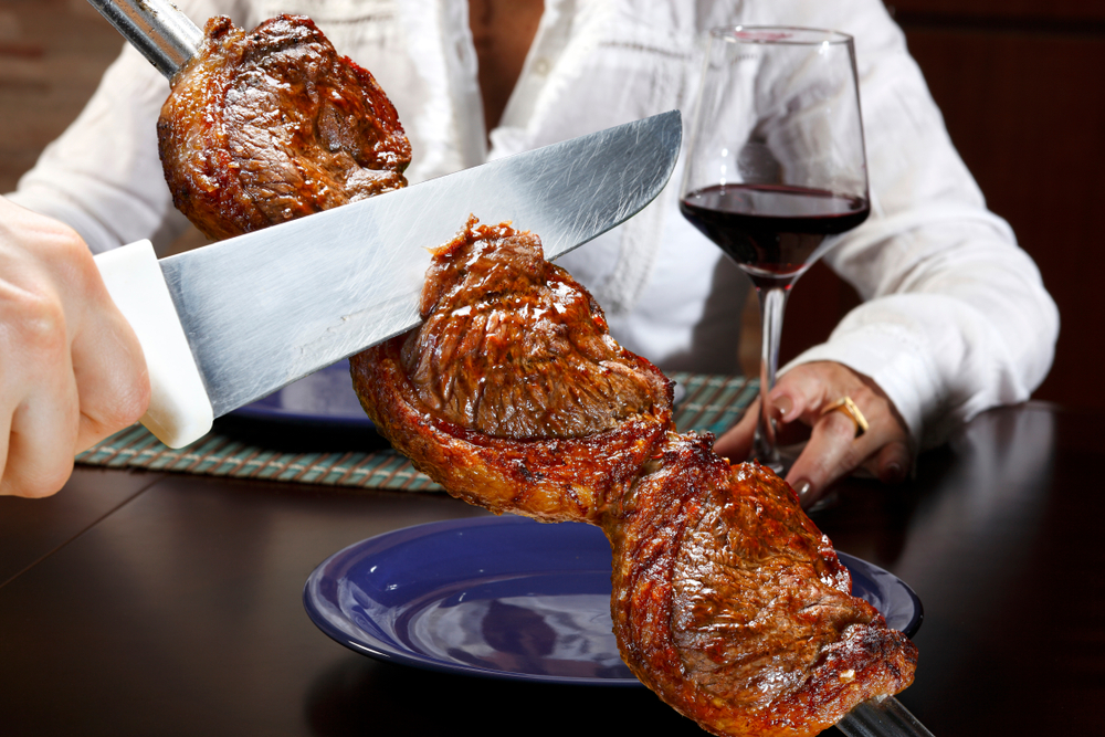 A knife slices meat off a metal rod, in the style of Brazilian steakhouses, to be served to a woman sitting at a table, with a glass of red wine.