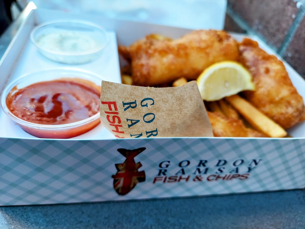 A box of fish and chips with two sauces fit into the box and Gordon Ramsay's label on the side and on the napkin.