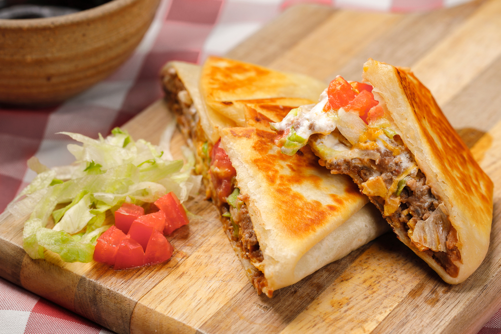 A crunchy "wrap", filled with meat, cheese, and veggies on a wooden board with small slices of tomato and greens next to it, similar to the vegan version served at Leguminati, one of the best restaurants in Orlando for vegan food.