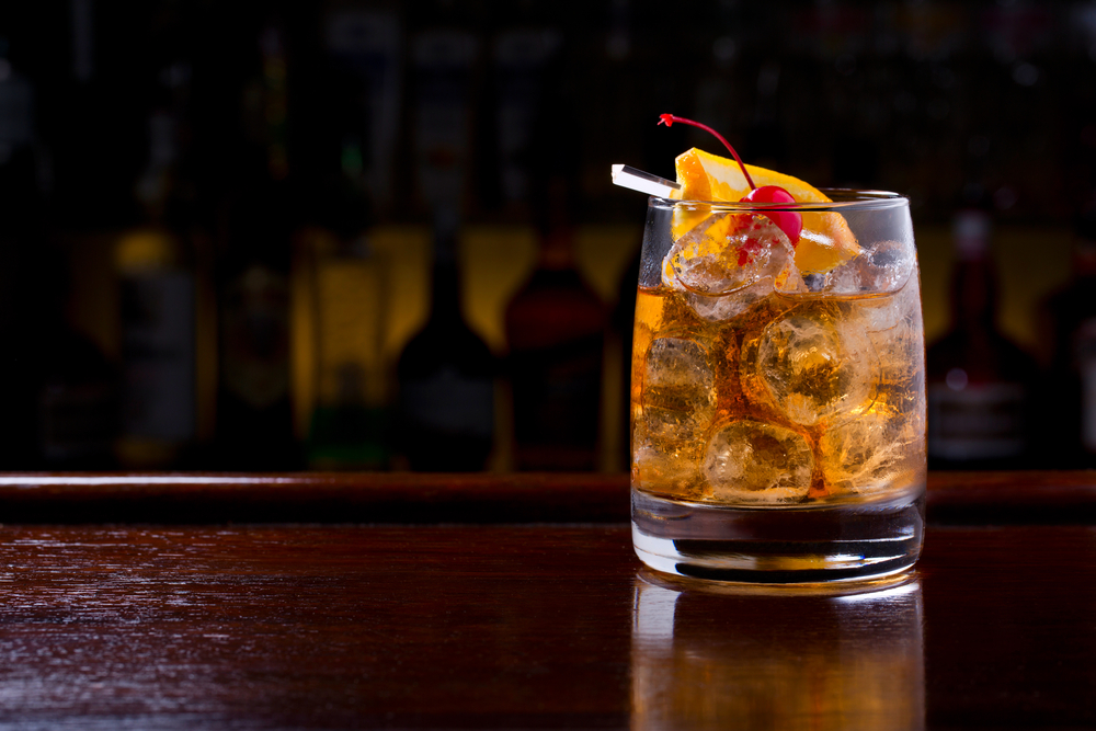 An old fashioned cocktail, with a cherry and orange peel, sits on a dark wooden bar, like that in the Island Hotel in Cedar Key, FL.