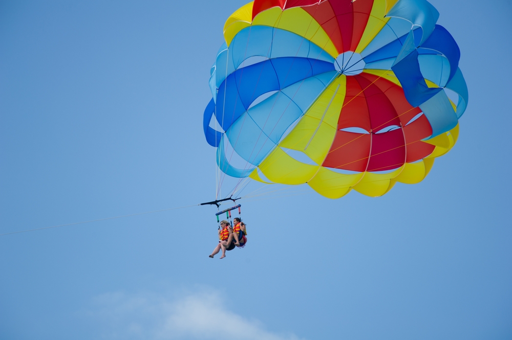 Two people go parasailing (one of the best things to do in Miami for adults), with a bright, multi-colored parasail against a blue sky.