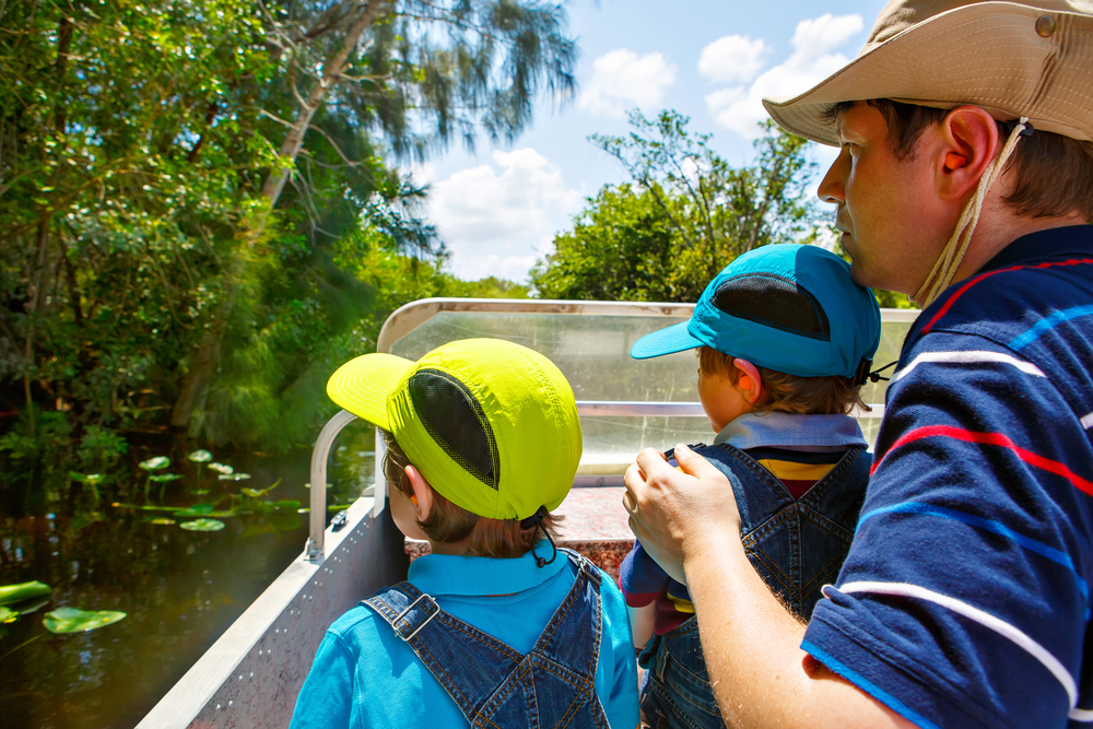 A father hovers over two sons in ball caps as they over look the edge of their airboat and into the waters and vegetation of the everglades. Peering for gators, these are some of the best airboat tours near Orlando FL. 