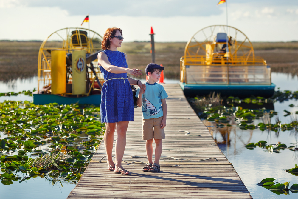 A woman and her young son walk a boardwalk that leads to two airboats. Vegetation and water surrounds them at one of the best airboat tours near Orlando. 