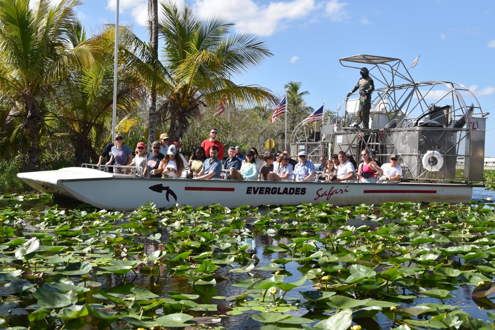 A long airboat with a guide on the back propels a bunch of people forward through the thick vegetation that floats on top of the water. 