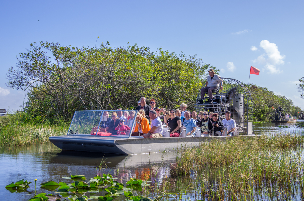 A bunch of adults are enjoying Orlando for Adults by taking a airboat tour of the everglades. They are crammed in the boat with a guide in the back, and the marsh surrounds them.