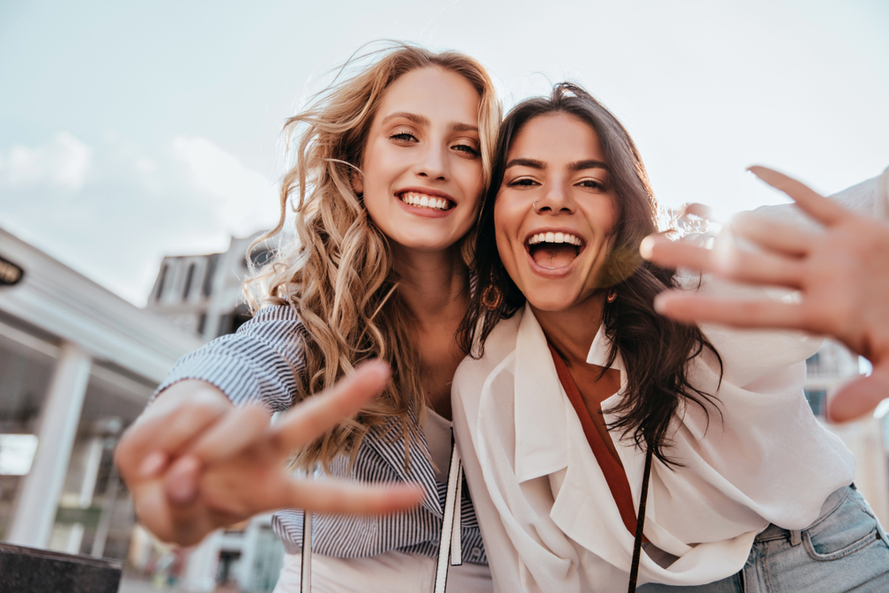 Two friends smile for the camera in front of a hotel, waving and throwing a peace sign at the camera lens.