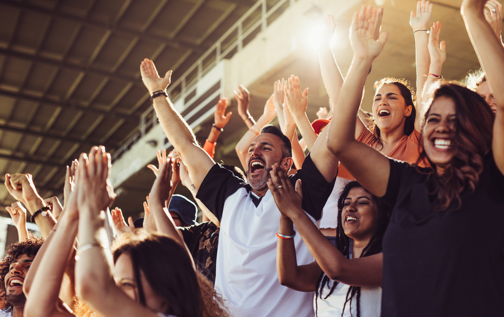 A series of adults in the crowd are out of their seats and cheering with their arms up in the air at this sports game.