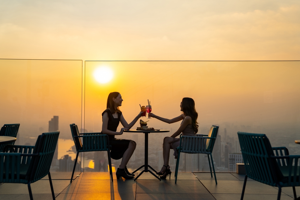 Two friends cheers their drinks on a rooftop bar with the sun setting in the background. 