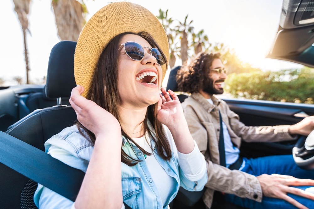 Happy couple driving convertible car enjoying summer vacation in Florida. The girl is laughing and wearing a hat. 