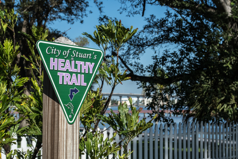 A triangular green sign on a post that states "City of Stuart's Heathy Trail" in front of trees and the water