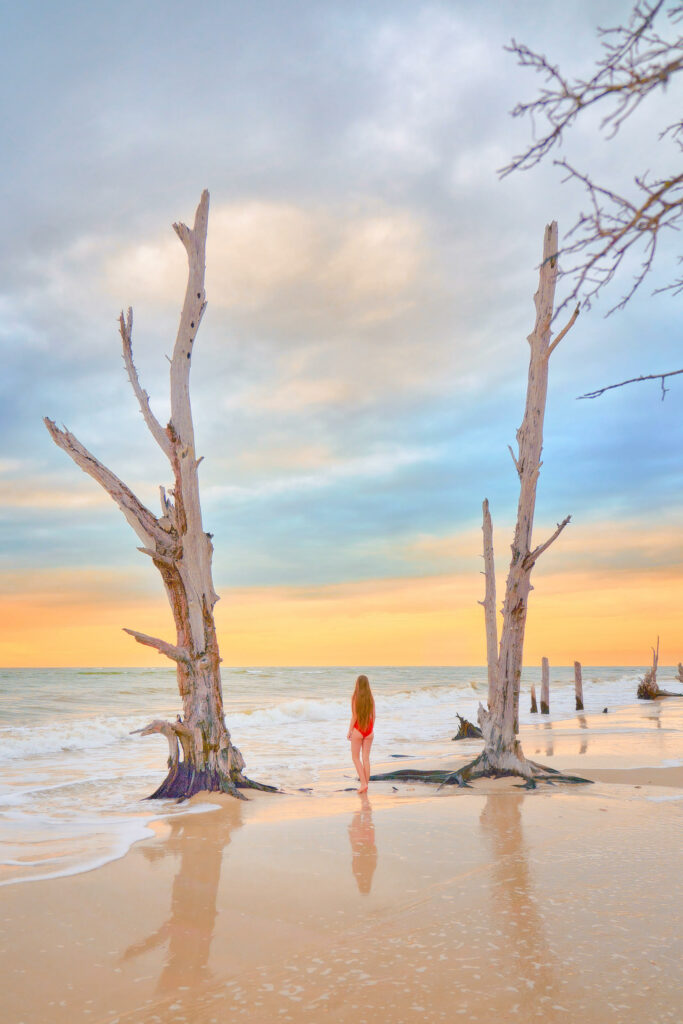A woman in a red swimsuit stands on a beach at Lovers Key with driftwood trees at sunset.