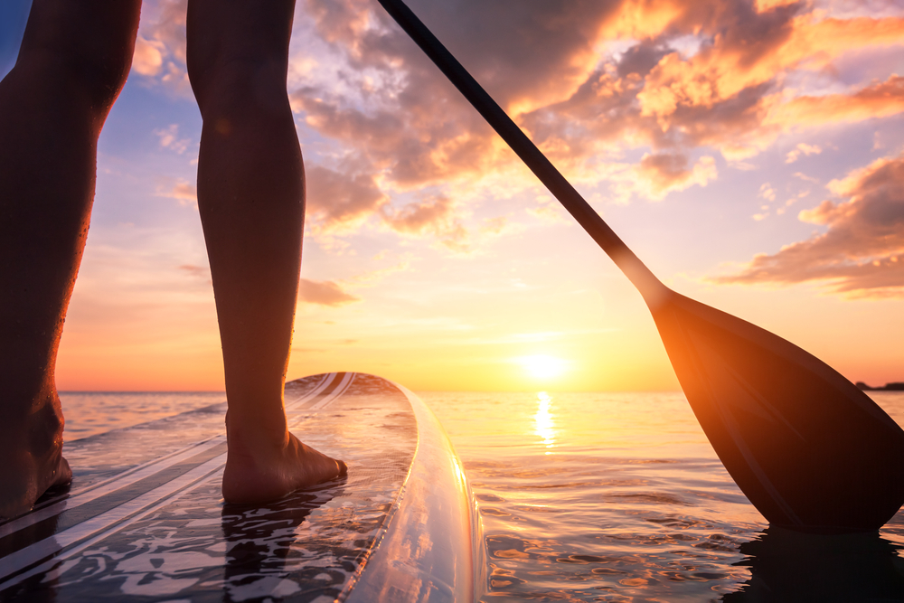 A closeup of two feet on a stand up paddle board, with a paddle dipped in water, at sunset.