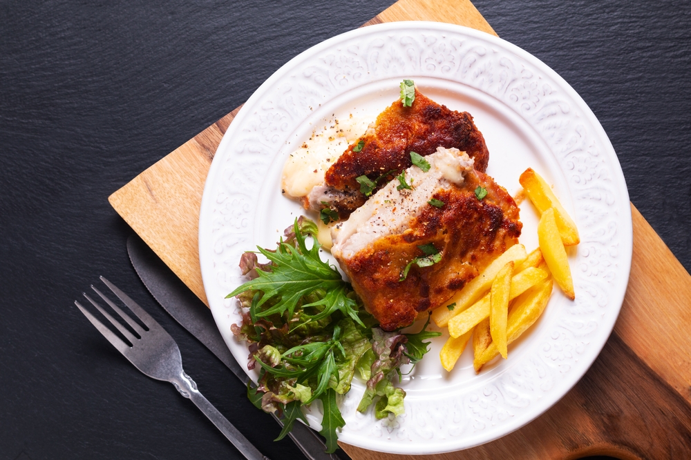 Chicken Cordon Blue sits on a white plate with a fork next to it: the entree also creatures a side salad and French fries! 