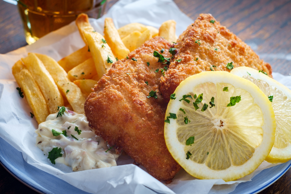A classic fare of fried fish and chips sits on a plate with extra lemon and Tartar sauce on the side.