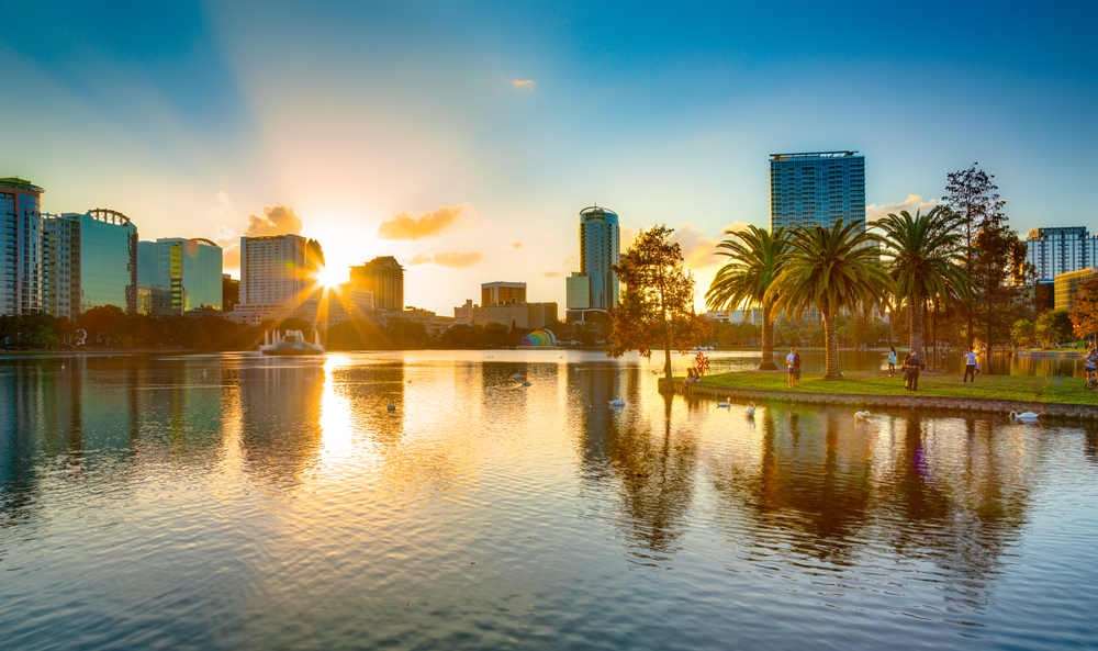 In central Florida there are big cities growing: places like Orlando make living in Florida popular. This photo shows the city over a giant lake at sunset with people walking around it. 