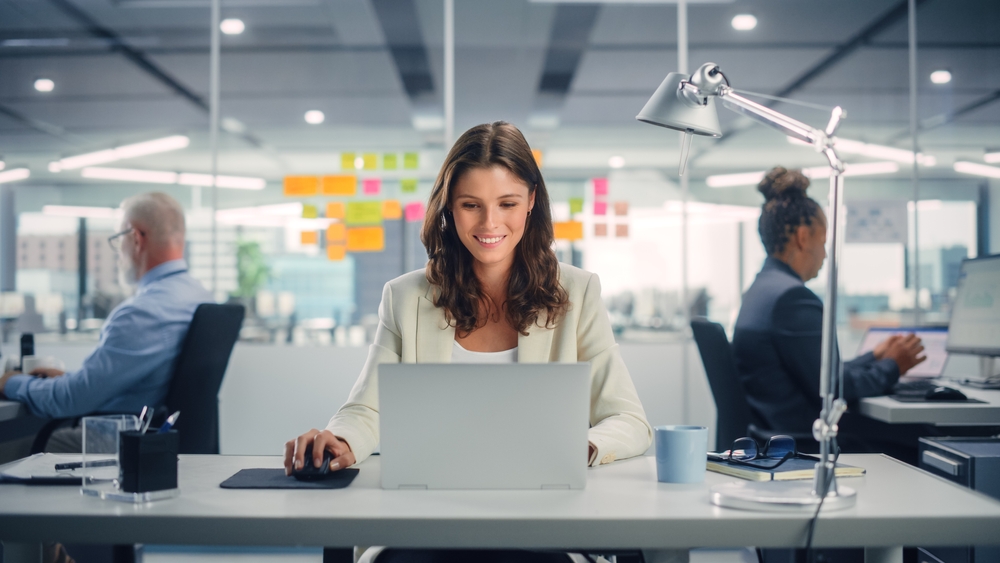 A woman sits at her laptop at a new job, and two people behind her keep working. There is a great growing job market in FL. 