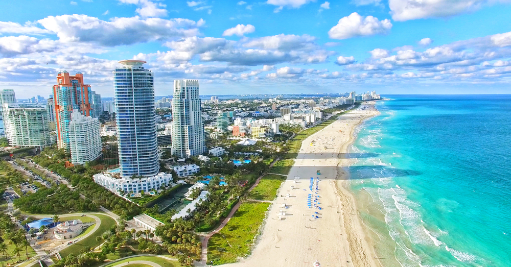 Looking to live in Miami? This coastal city is stunning, as seen in this photo with its blue waters, tan sand, and great city markers, but there are some cons to it too. 