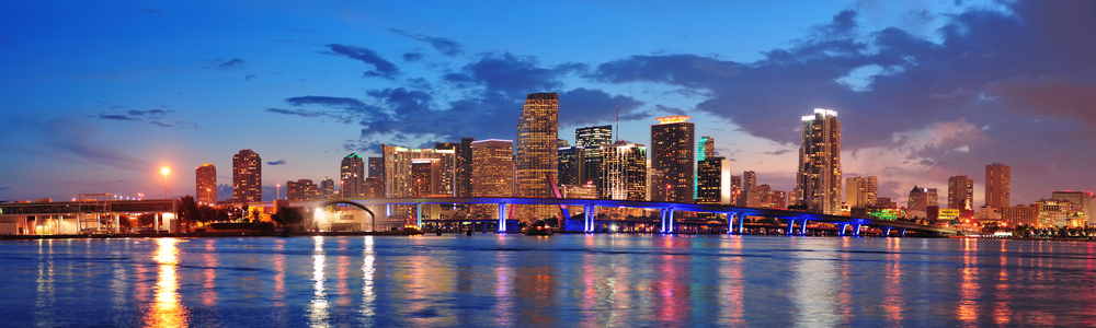 The cityscape of Miami at night features tall buildings that glow with lights and reflect off the water. 