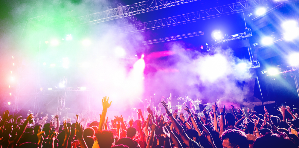 Tons of people gather together and jump and and down to the beat of music at this nighttime event and festival in Miami. Smoke fills the air and bright lights cast shadows over the crowd. 