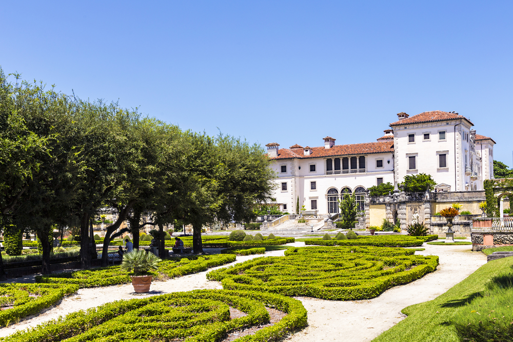 a big European style building with green gardens in the front. there are also olive trees up against the big white building with a blue sky 