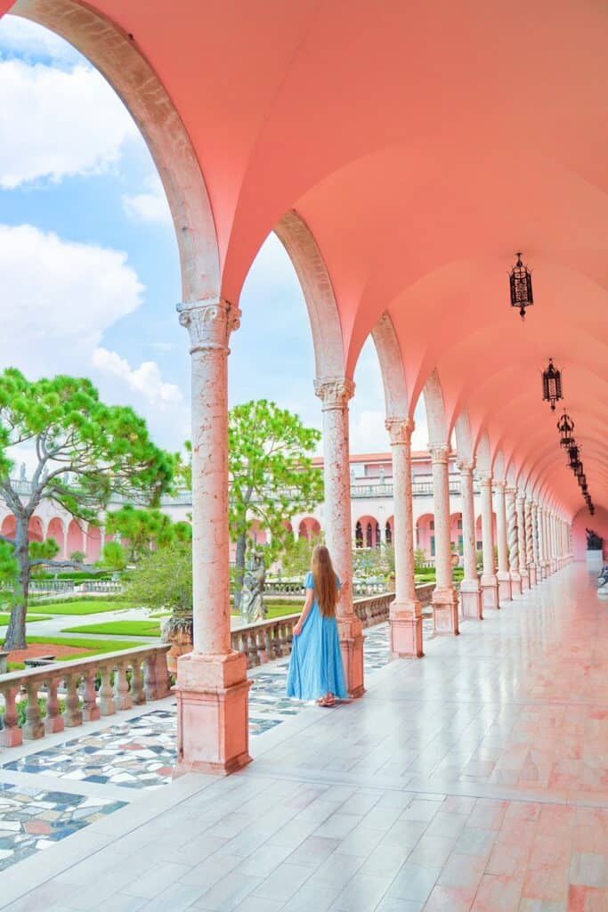 Victoria standing in front of a beautiful garden inside of a museum with pink walls and European architecture. she is wearing a blue dress with her long hair down 