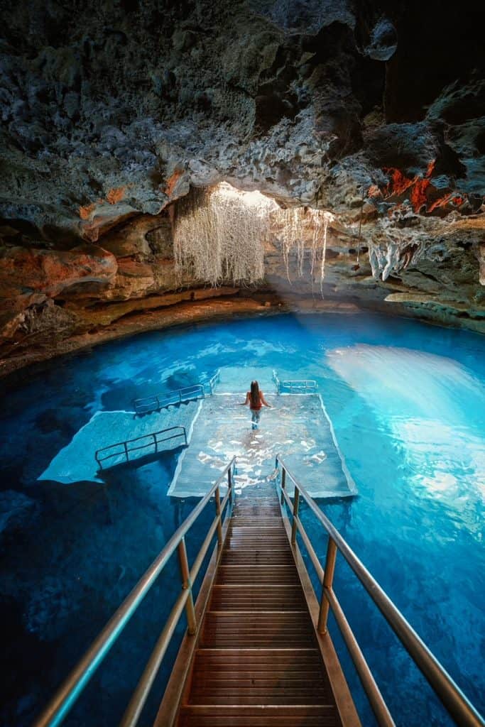 woman standing with hair down in red bathing suit in a walk down onto a platform ready for a dive in beautiful bright blue waters with a big ray of light shining from an above ground opening in the cave