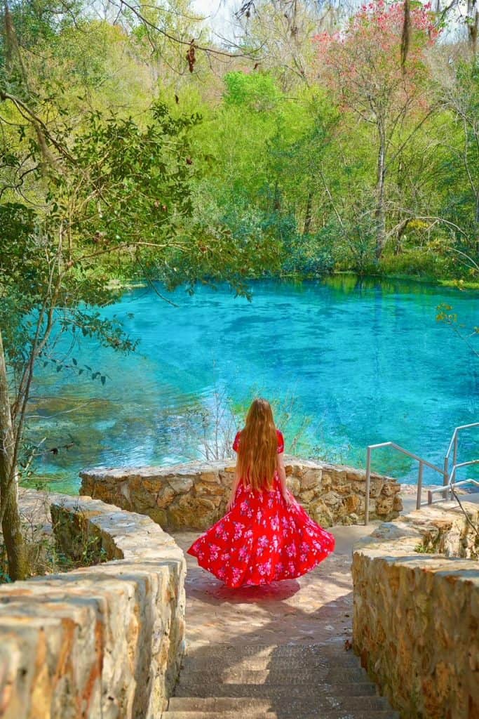 woman in red floral dress stands on rocky path down to water staring out over the bright blue waters and bright green trees 
