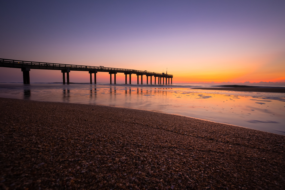 During a sunrise in Saint Augustine, the sun glows orange over a pier and the shelly texture of this coastline. 