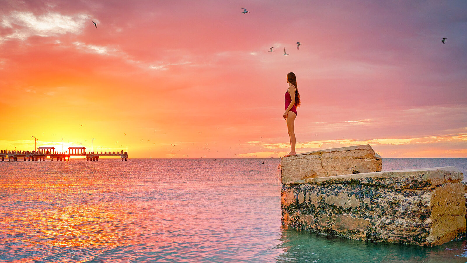 A woman in a red bathing suits stands atop an old piece of a fort on a coastline, offering perspective of one of the most beautiful sunsets in Florida.